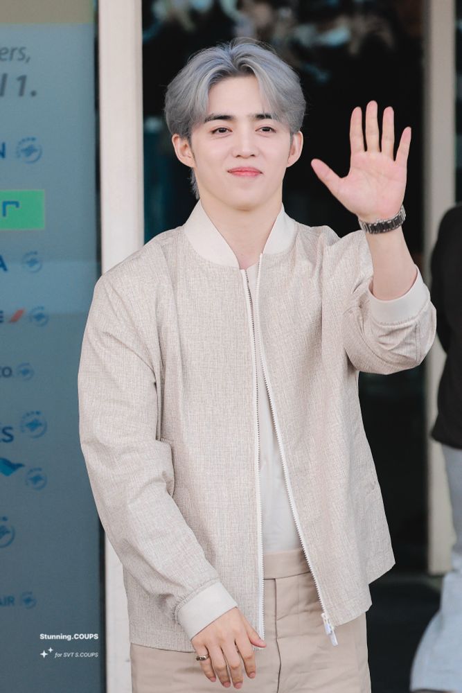 scoups wearing neutral on neutral. his hair is a blue grey. he is wearing a gold bracelet and silver watch. he is waving at a camera and smiling. his team ring is visible.