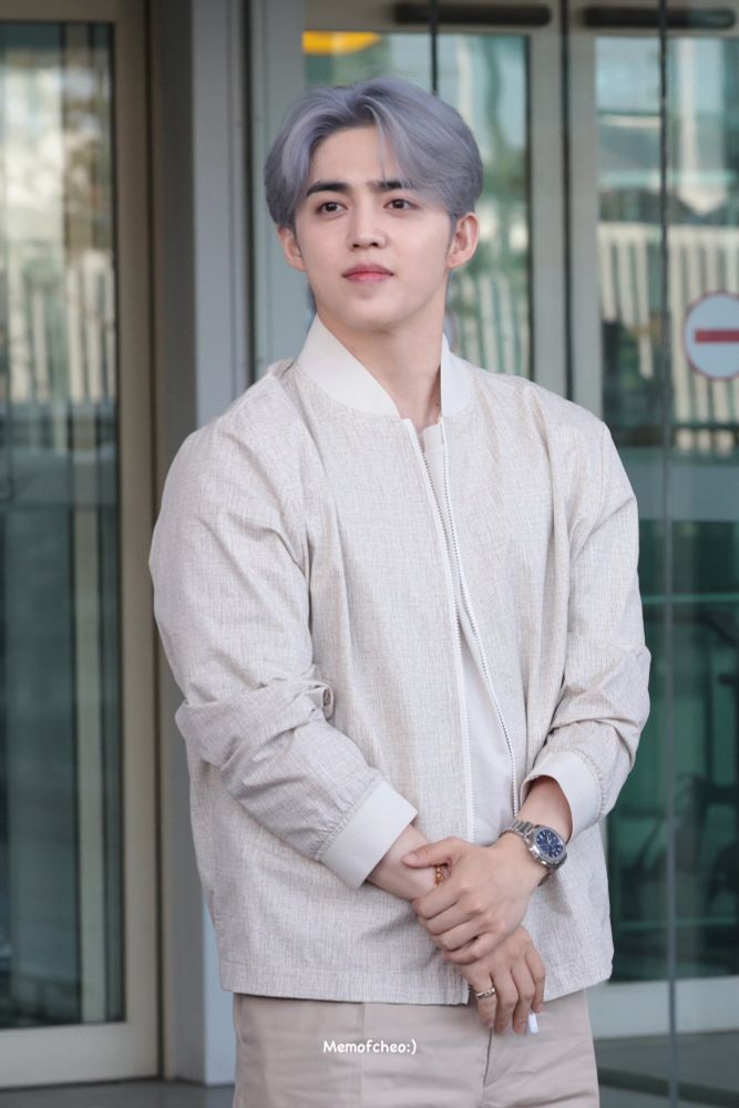 scoups wearing neutral on neutral. his hair is a blue grey. his hands are crossed, hand over wrist. he is turned to the side, looking concerned.