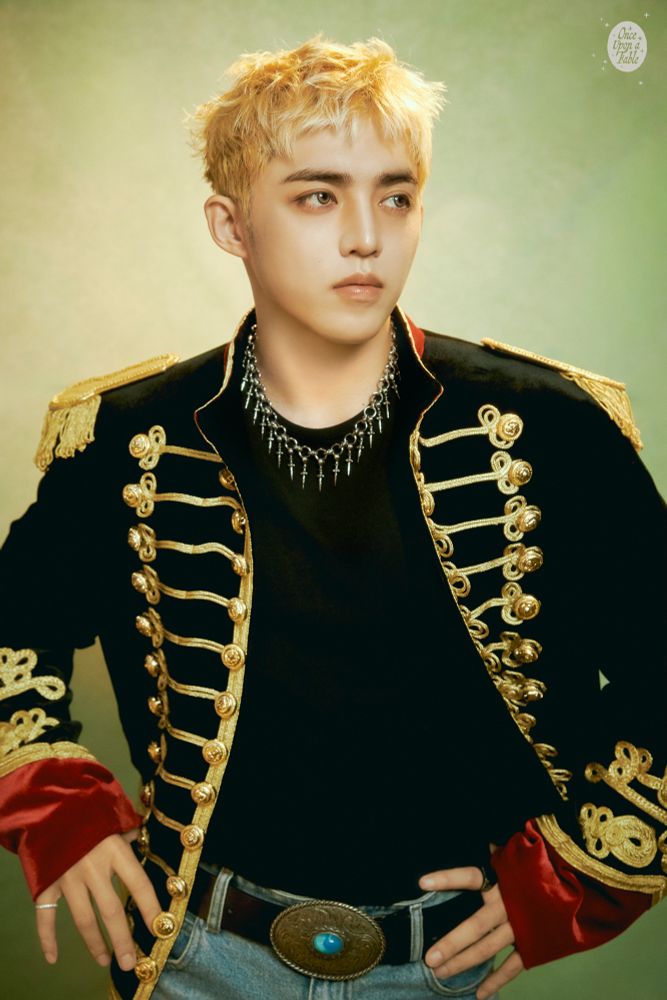 scoups in an open black uniform top with gold detail and red at the end of the sleeves, jeans and a belt with a large belt buckle. he is blond, looking away from the camera. 