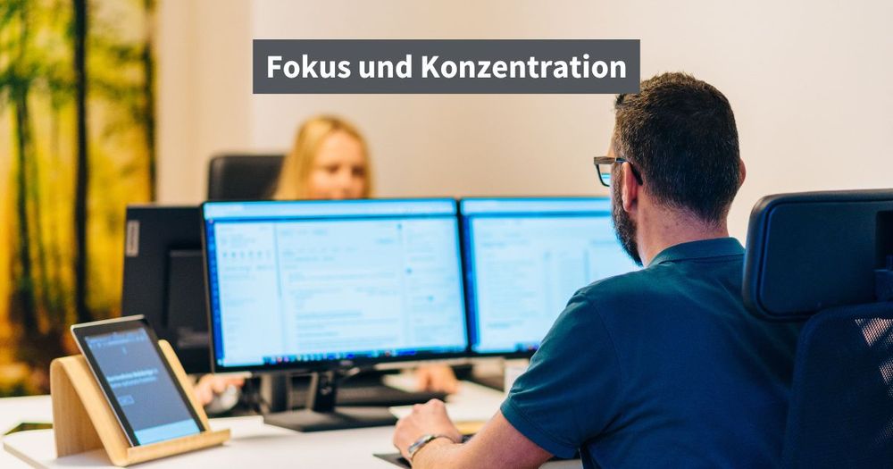 „Oliver & Katharina sitzt an ihren Schreibtischen mit zwei Monitoren gegenüber und arbeiten konzentriert. Auf dem Bild steht der Text ‚Fokus und Konzentration‘.“