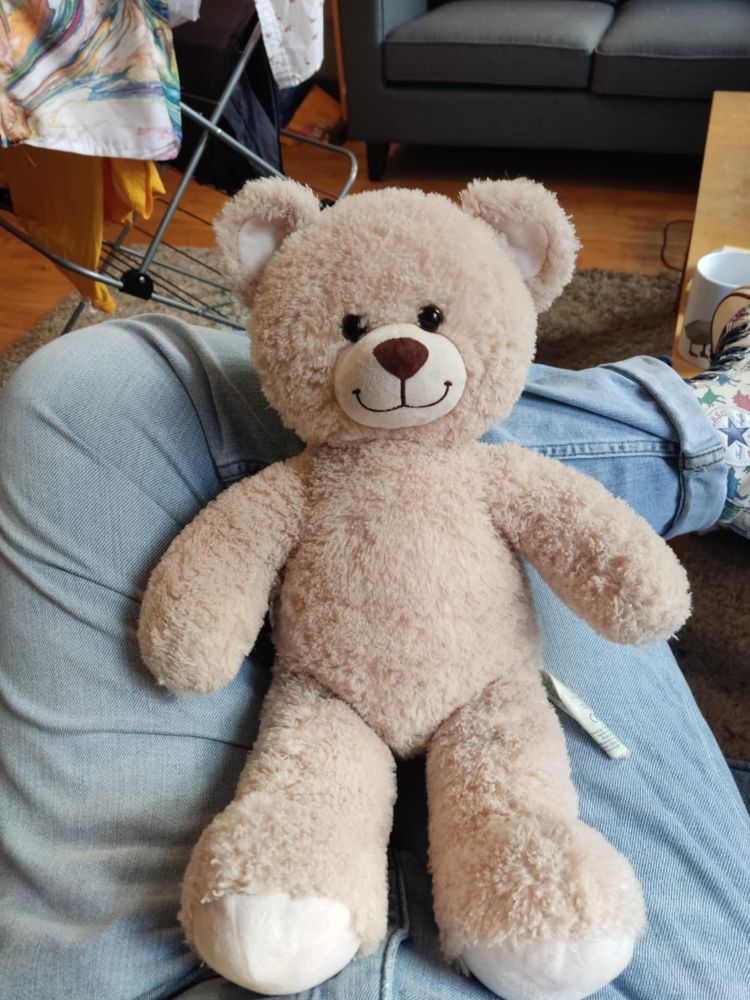 A beige Build-A-Bear teddy with a smiley face sitting on the lap of someone wearing pale blue jeans.
