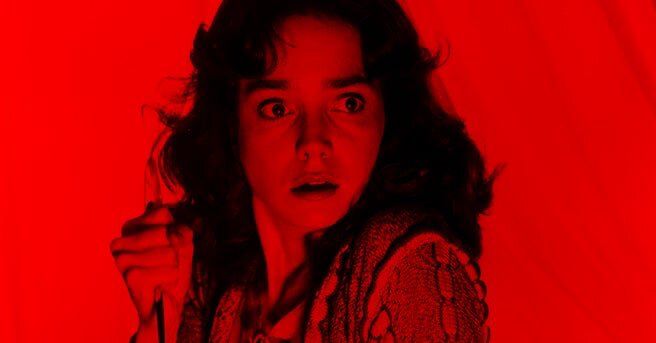 A close up on the face of a frightened young woman with dark hair clutching a knife close to her shoulder. The whole frame is a saturated red colour. It is a still from the 1977 Italian Giallo horror movie Suspiria.