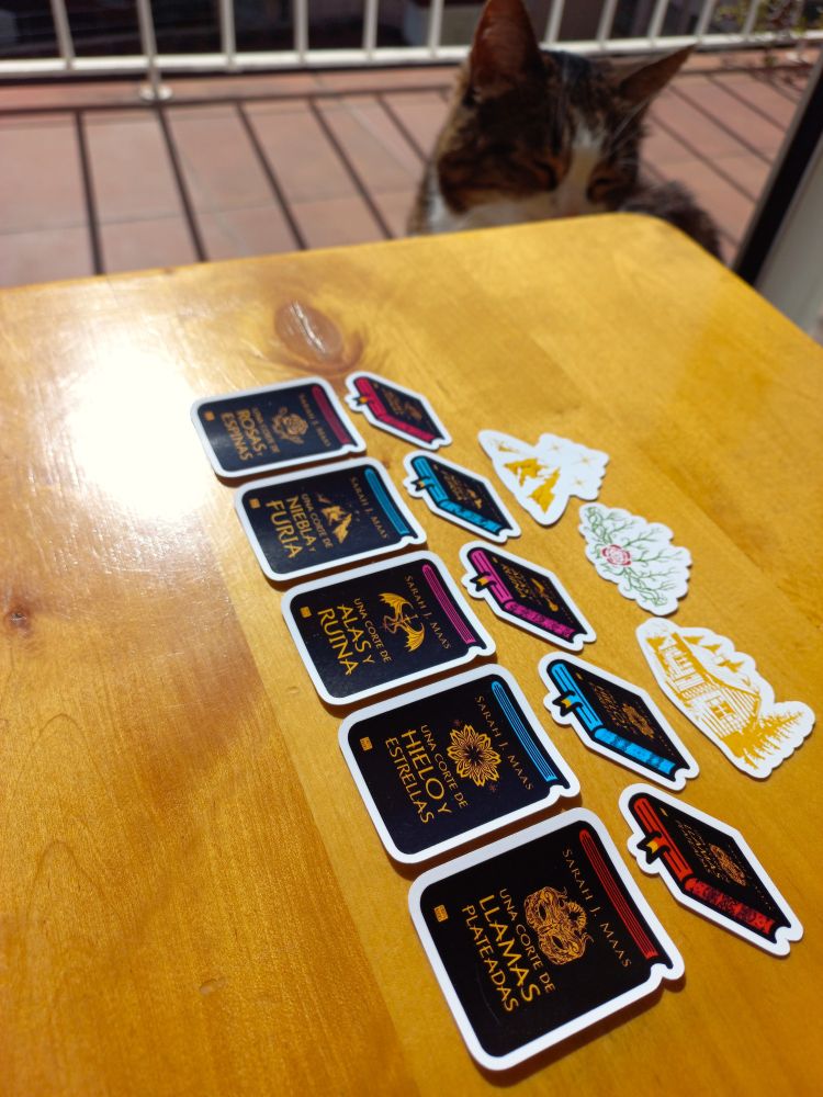 Photo of a small wooden table in the foreground on which there are some stickers of the ACOTAR book saga, in the background you can see the bars of a balcony and there is a cat next to the table with only her head showing.