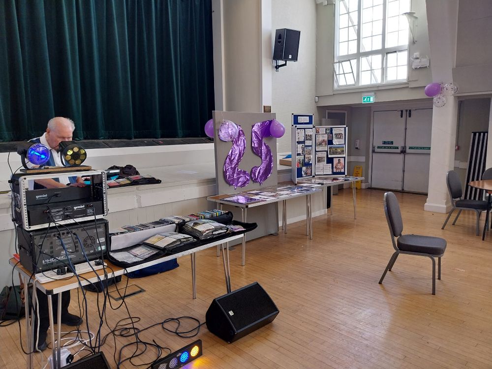 A man sets up DJ equipment on stage in a hall. A display with a large purple "25" and informational boards is nearby.