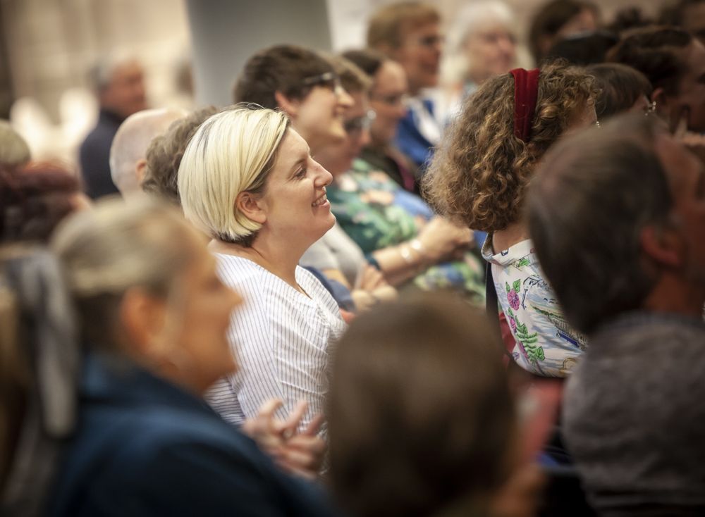The image shows a group of people seated as part of a crowd at an indoor event/meeting.