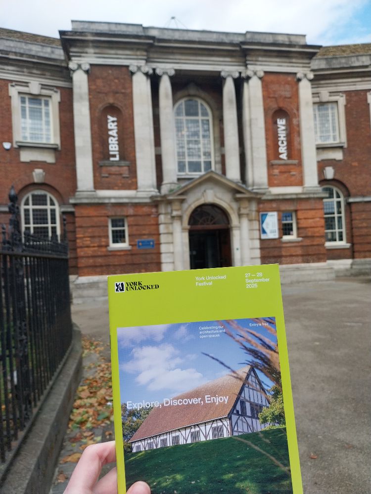 Person holds a York Unlocked Festival brochure in front of the historic building labeled "Library" and "Archive".