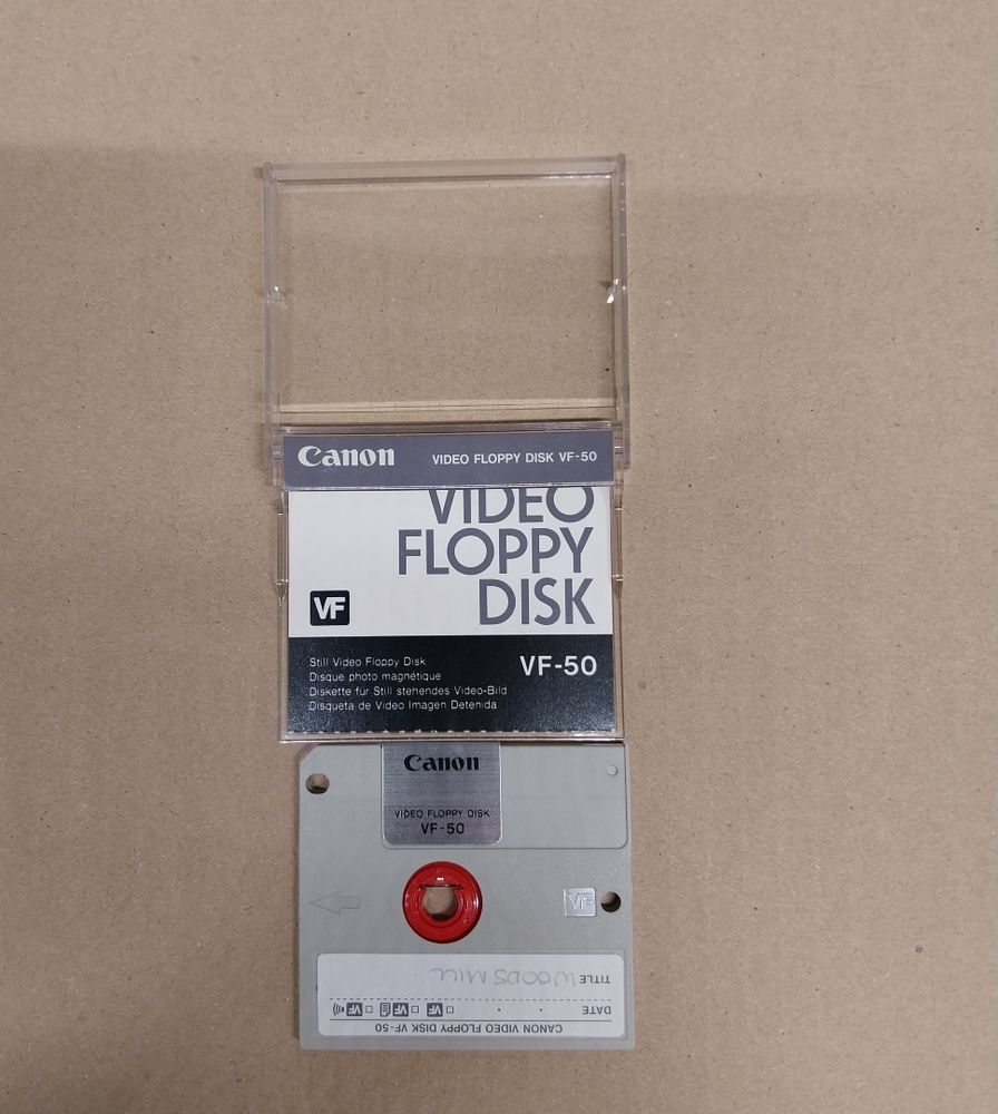 A small grey floppy disk, with a red circle in middle. The disk is in a plastic case with Canon branding on the top and 'Video Floppy Disk VF-50". There is a label on the disk that is upside down that reads "Woods Mill". The case is opened and there is further branding with a black and white theme with some technical specs for the disk. 