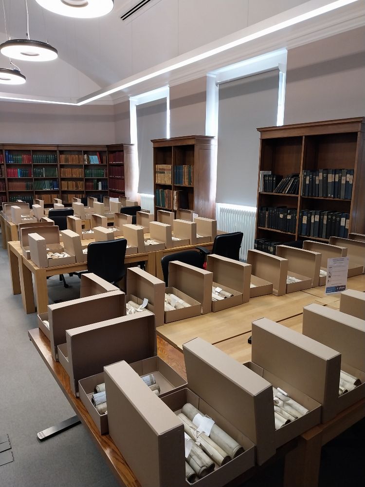 A room with tables lined with open archival boxes containing rolled documents, surrounded by bookshelves filled with books and ledgers.