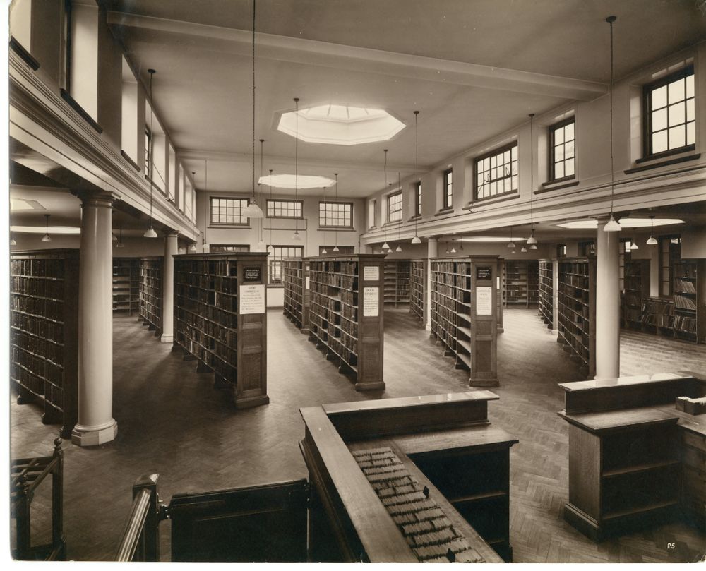A vintage library interior with tall bookshelves, large windows, and a central skylight, creating a well-lit, spacious reading area.