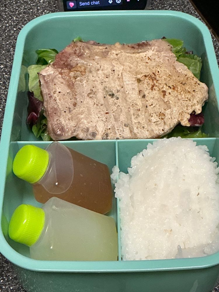 A porkchop rests on a bed of salad, one pocket has rice, the other has dressing and lemon juice in little bottles
