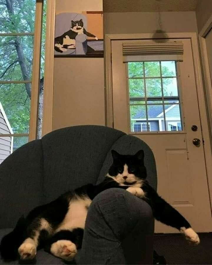 Photo of a cat sitting in a chair beneath a painting of the same cat sitting with the same pose 