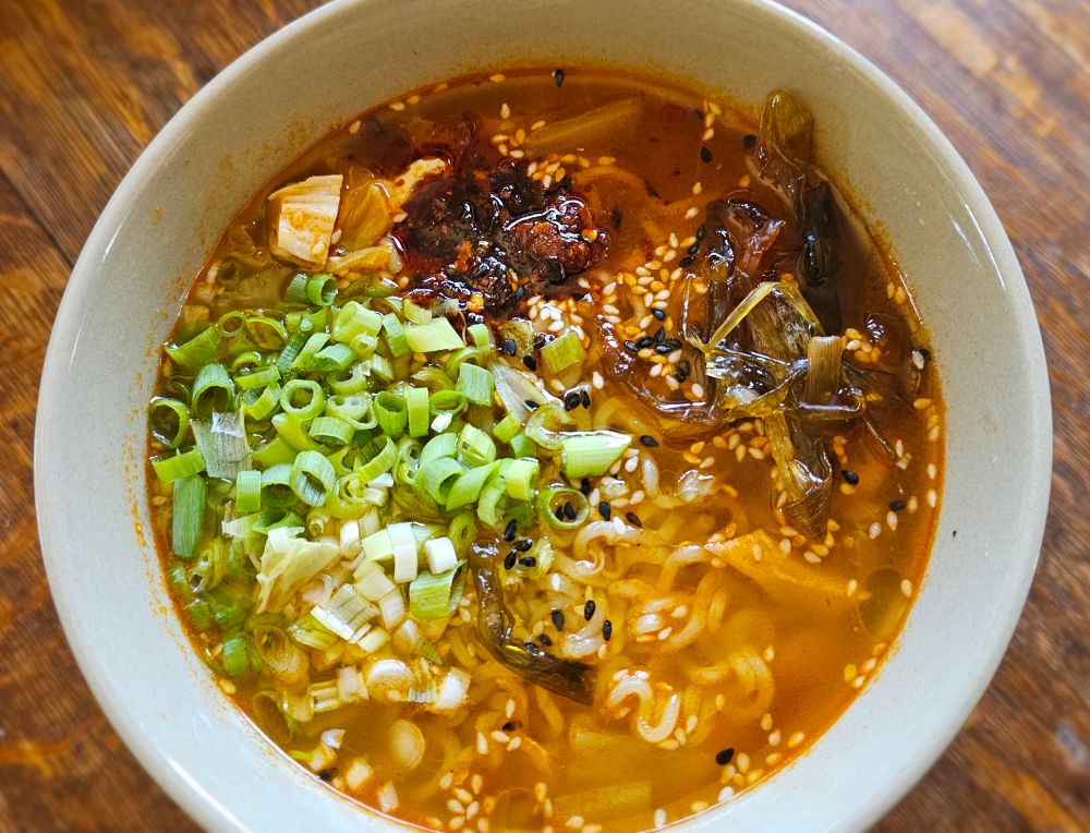 top view bowl of Samyang instant ramen with fresh scallions, fried scallions + oil, chicken, grated fresh ginger & garlic, sesame seeds, cabbage, & crispy chili flake 