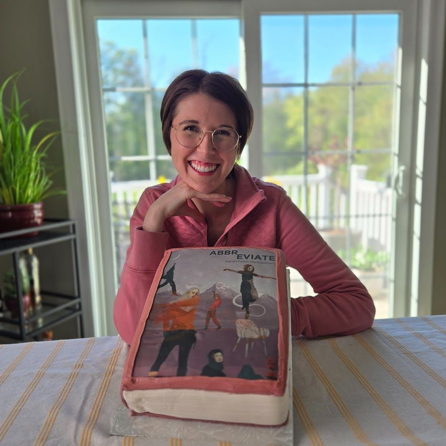 A pixie haired woman in gold round glasses and a pink shirt smiles while holding a large pink cake decorated like a book and featuring the cover of “Abbreviate.”