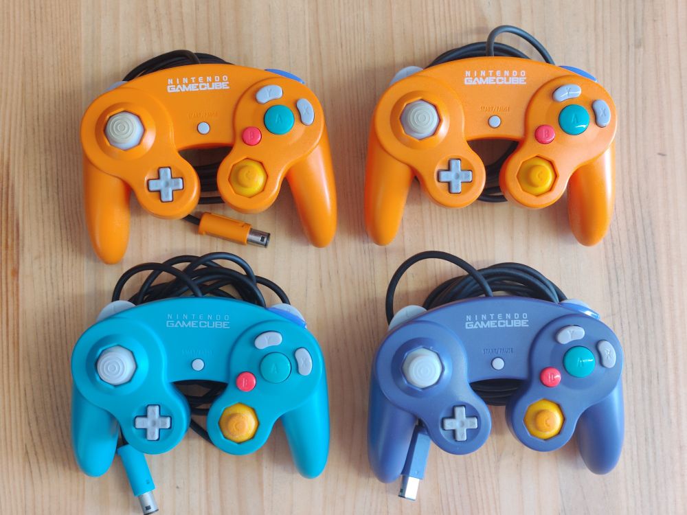 4 Nintendo GameCube controllers on a wooden background.
Top two are orange, bottom left is emerald blue, bottom right is purple.