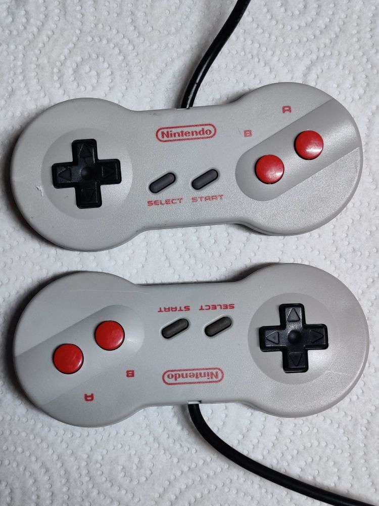 Two Nintendo Famicom "dogbone" controllers on a paper towel rotated 180° from each other, buttons facing toward the camera. Difference in color is barely visible.