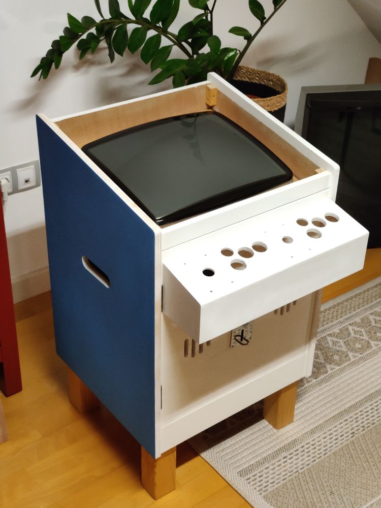 An invomplete blue and white Dagashiya arcade candy cab placed with two legs on a carpet and two on the wooden flooring.
In the background there is a potted plant and a B&O MX4002 tv.