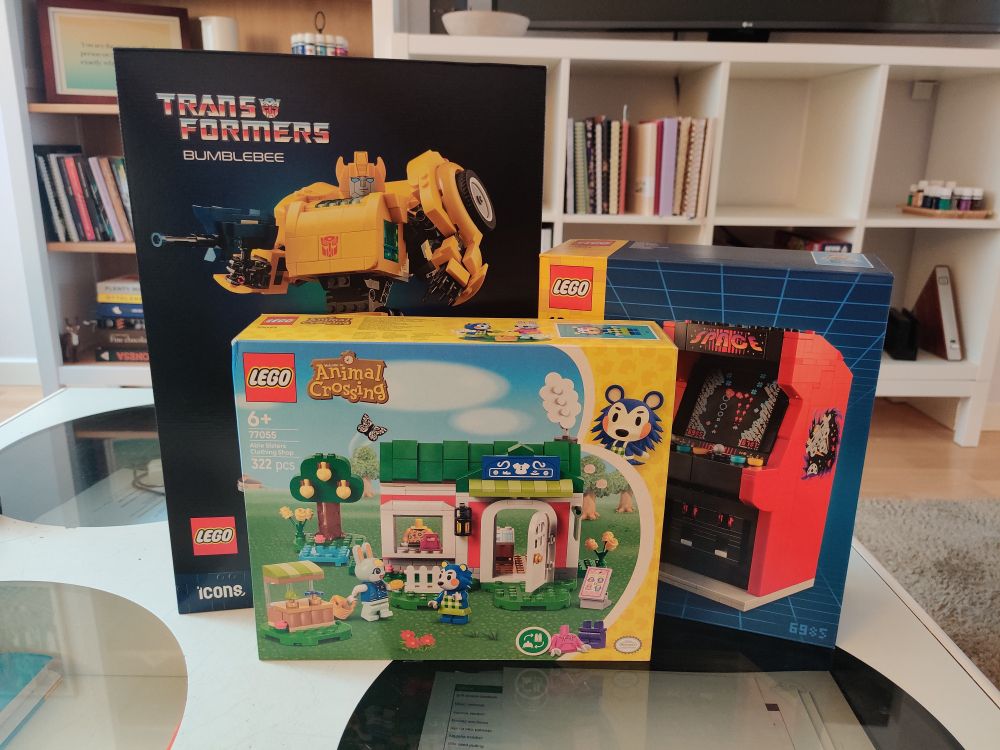 Three boxes of Lego (Bumblebee, Animal Crossing - Able Sisters, and Arcade machine) on a table with bookshelves on the background.