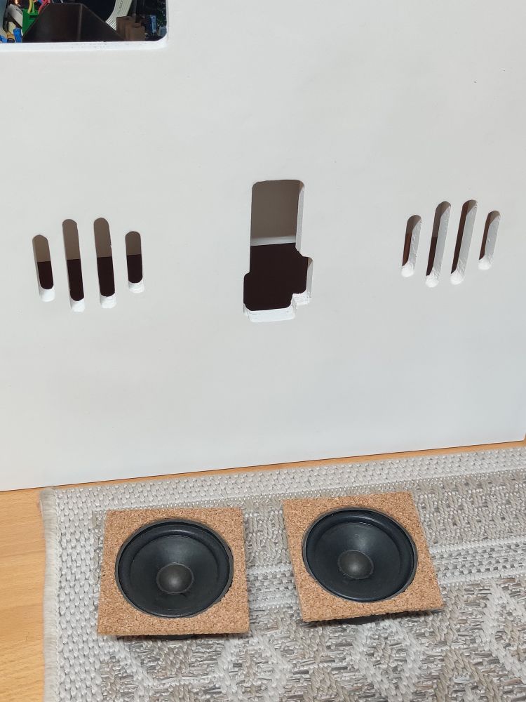 Two 3.3" speakers on a carpet with square cork damping mounted on them. Behind the speakers is the arcade door that they will be mounted on.