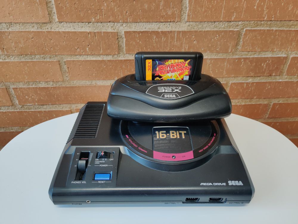Sega (Asia PAL) Mega Drive with (European) 32X plugged into it and (North American) Shadow Squadron plugged into the 32X on a white table in front of a brick wall.