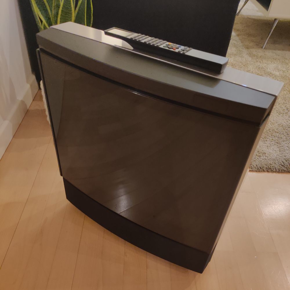 Bang and Olufsen MX 4002 TV with remote on top.
The TV is black with grey back (they came in several color combinations) is sitting on a laminate floor with a plant, carpet, and couch behind it.
