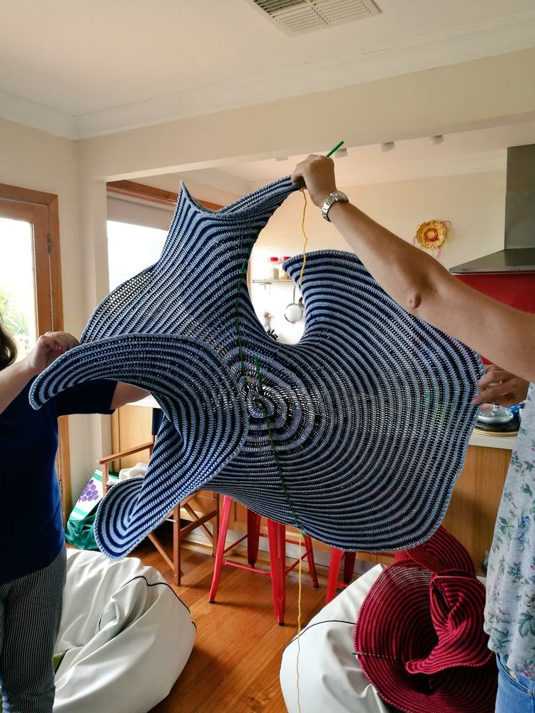 Blue crocheted surface being manipulated by two people whose arms and hands are visible.