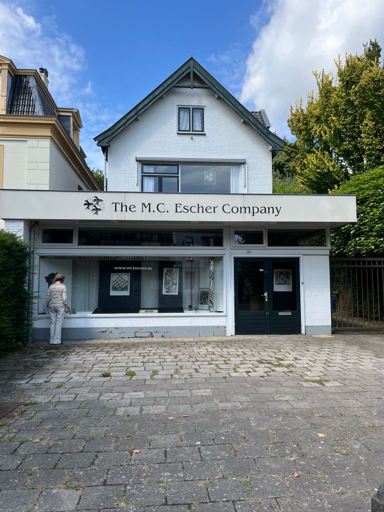 White shop with black writing and artwork of Escher displayed in the windows. 