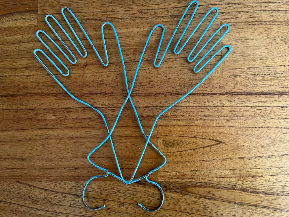 Two pieces of blue wire each bent into the shape of a hand with five fingers and a hook to use as a hanger for gloves.