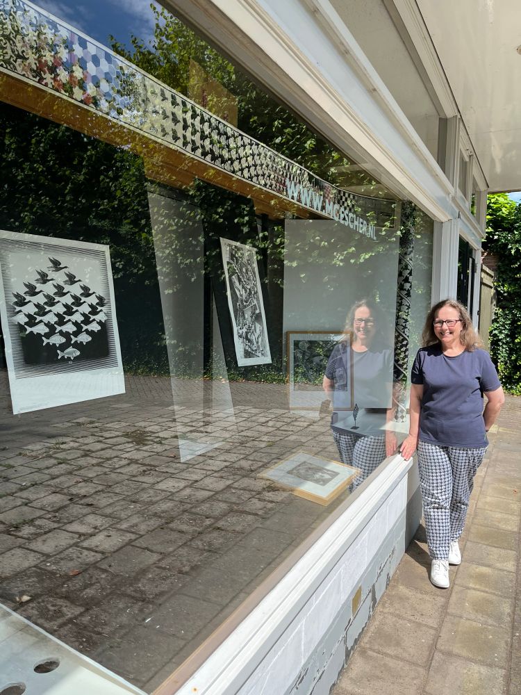 Woman standing beside a glass window. Her reflection is visible beside her. The window displays works of Escher. 