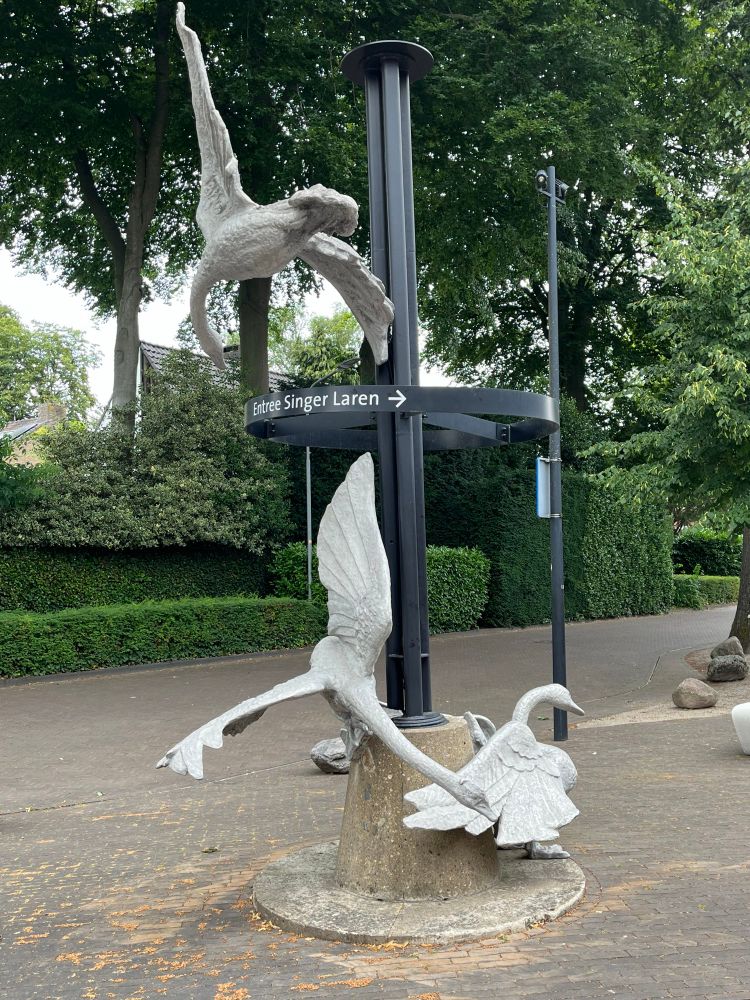 The three swans made of white metal around a black pole outdoors. The signage says Singer Laren. 