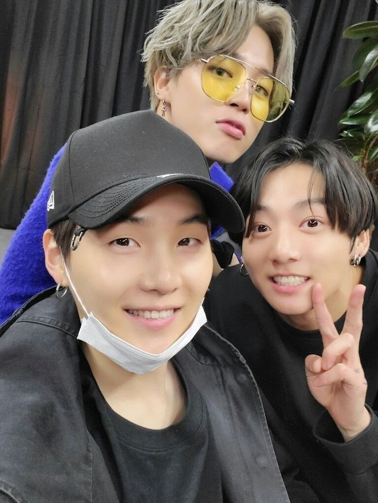 A picture of Jimin, Jungkook and Yoongi. The latter is wearing all black (shirt, jacket and cap) and a white face mask under the chin, JK is wearing a black sweater, Jimin a blue one with yellow tinted sunglasses, and his hair is light gray. They're sitting next to each other.