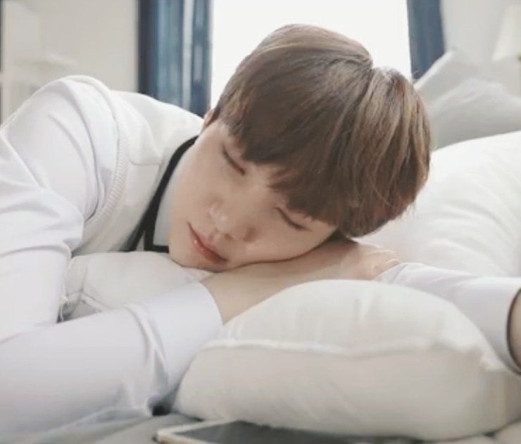 Yoongi laying down with a pillow, with his eyes closed. He's wearing white and has brown hair.