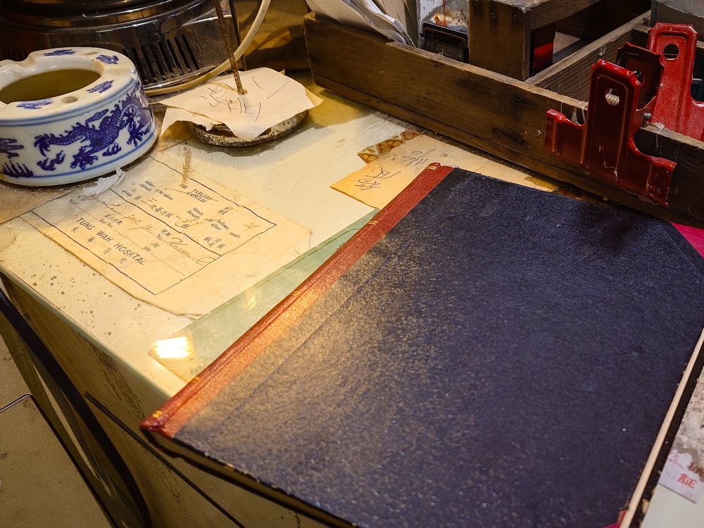 映画『トワイライト・ウォリアーズ』九龍城寨公園の展示写真。七記冰室のカウンターの上。黒い表紙の帳簿や龍柄の陶器の灰皿が置かれ、様々な書類がある。