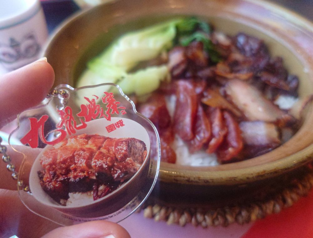 九龍城寨之圍城の叉焼飯アクリルキーホルダーを左手に持ち、背青梗菜、中華ソーセージ、腊肉が乗った土鍋ご飯を背景にした写真。