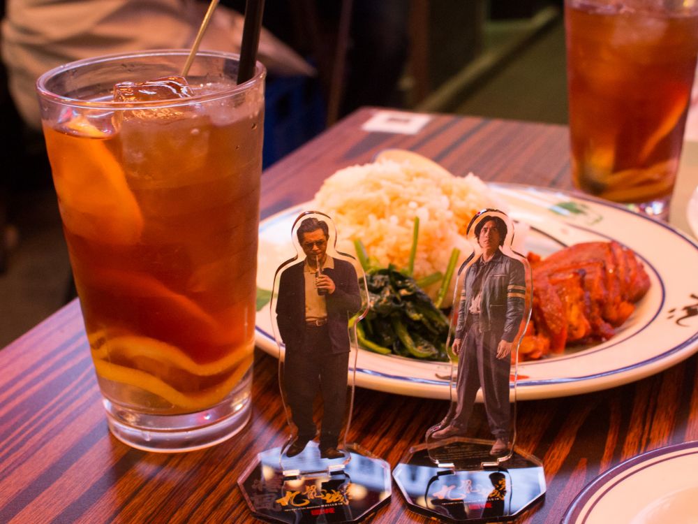 机の上、斜俯瞰写真。アイスレモンティーと鶏柄の丸皿に乗った叉焼飯、映画『トワイライト・ウォリアーズ』の龍捲風と信一の円盤特典アクスタ。