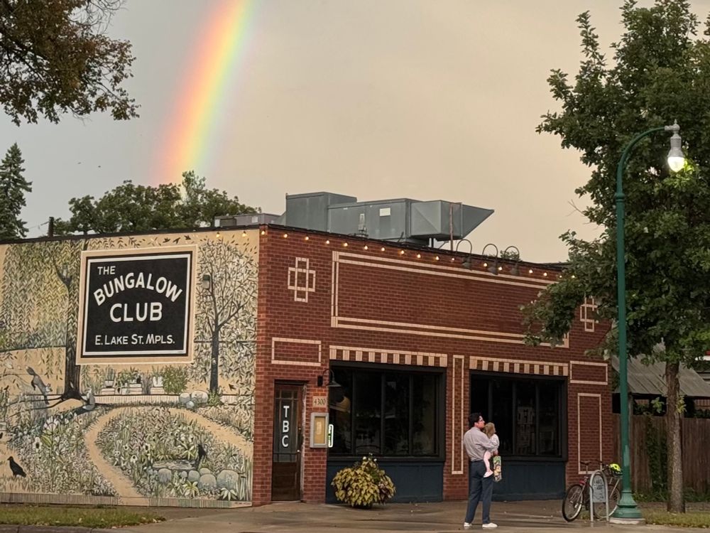 A rainbow in a gray sky appears to end behind trees. The main part of the photograph features a brick building with a sign reading “The Bungalow Club” and a mural on the side. A child and her dada are hugging each other in front of the building, waiting for the rest of their family to come outside.
