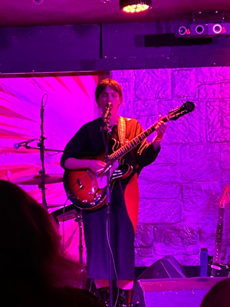 Dana stands on stage alone, singing into a microphone. She is playing a red guitar resembling a Gibson ES 355