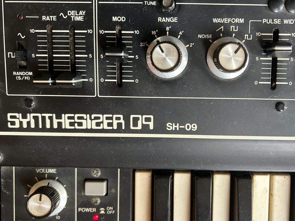 A close up of a section of a Roland SH09 analog synthesiser. Showing some keys, volume switch, range and waveform knobs and lots of switches and sliders.