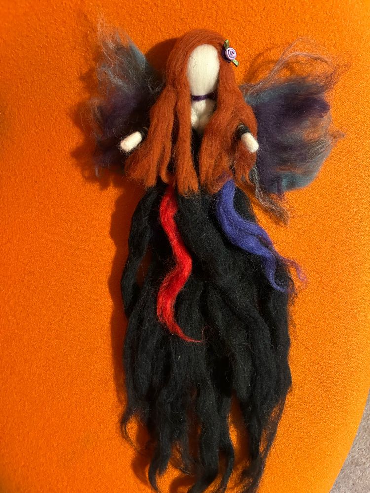 The image shows a red headed felted faerie with green, purple and grey wings. She has a purple choker and lots of cleavage on show. Her dress is black and flowing with red and purple accents. She has a purple flower in her hair and has been made using the felting technique