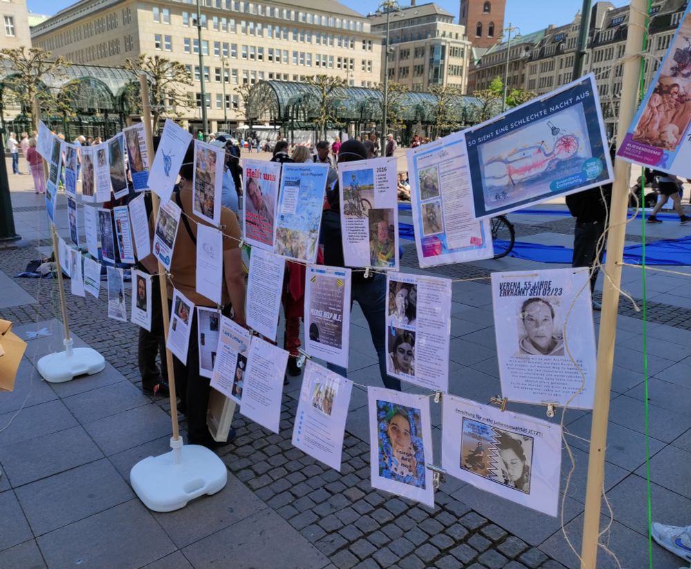 Hamburg, Rathausmarkt, heute, 11.05., auf der #Liegenddemo für von MECFS betroffene Menschen. Auf dem Foto sind viele Porträts von betroffenen Menschen zu sehen, die, aufgereiht zwischen Sonnenschirmständern und Holzstielen, als laminierte Fotos und Texte in drei Reihen untereinander an Bindfäden im Wind schwingen.