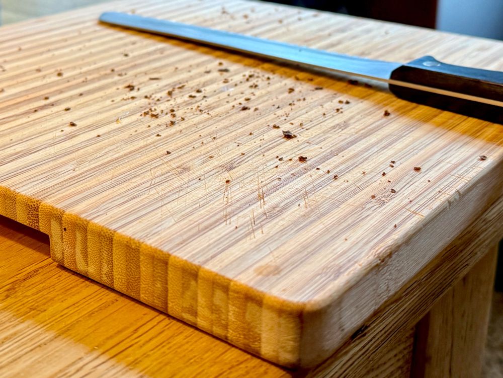 Ein Holzschneidebrett mit leichten Kratzspuren ist auf einem Holztisch zu sehen. Einige Krümel liegen verstreut auf dem Brett, und ein großes Messer mit einem schwarzen Griff liegt darauf. Die dominierenden Farben sind helle Holztöne. 