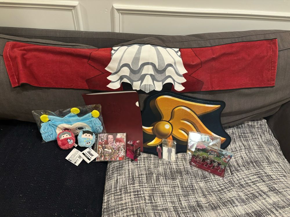 A picture of the merch order spread out on a grey couch with blue and grey blankets covering the cushions. It consists of:

-A long scarf-like towel spread out in the background, patterned to look like Miles Edgeworth’s jacket and cravat.
-A Blue Badger terry cloth headband.
-Two small plush keychains, one of Miles Edgeworth and one of Phoenix Wright, made to look as if the characters are stuffed inside small round sleeping bags. Miles’ is red and features a small design of the Steel Samurai; Phoenix’s is blue and features the Blue Badger.
-A red leatherette document folder with a portrait of Miles Edgeworth and his Japanese name, Reiji Mitsurugi, in a banner beneath it.
-The Ace Attorney Investigations Collection soundtrack CD.
-A standee featuring Miles putting on his jacket.
-A plush mat about a foot across that features the Yatagarasu pin design.
-Two keychains shaped like Miles’ and Phoenix’s suits.
-A standee featuring the game sprites of several Investigations characters against the background of the Grand Tower top floor.