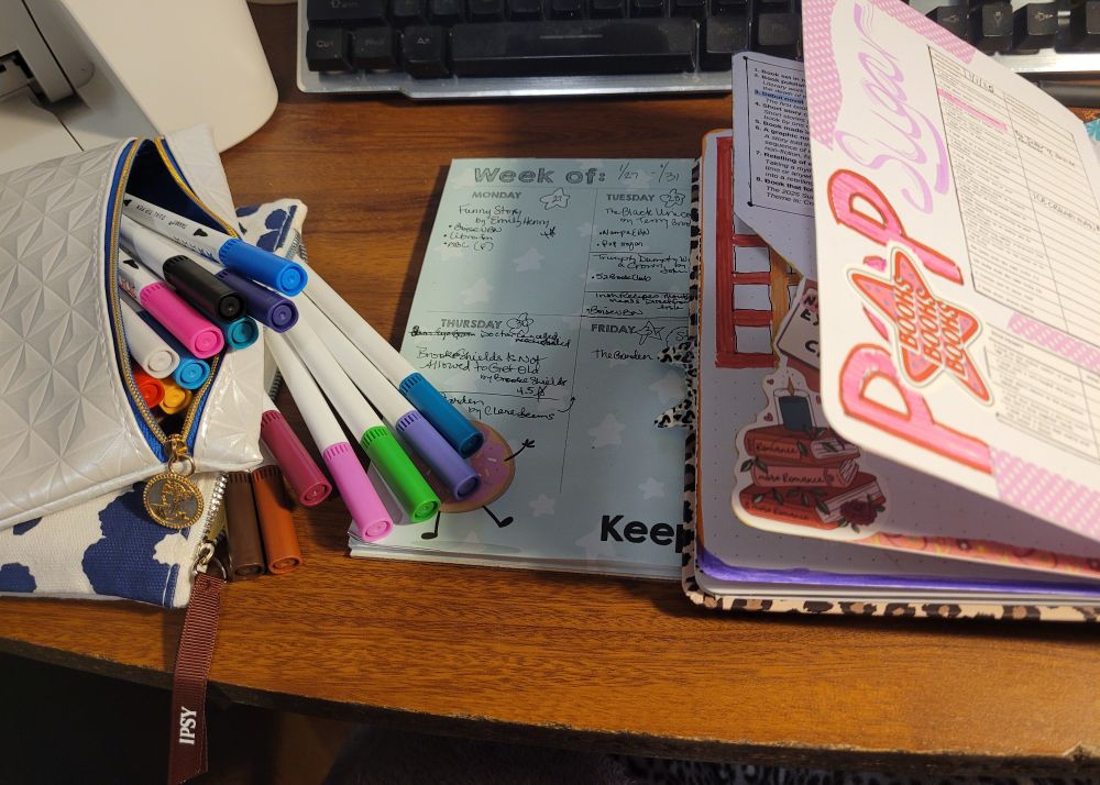 Brush pens, reading journals, & Ipsy bags on a wood pattern desktop. Keyboard in view.