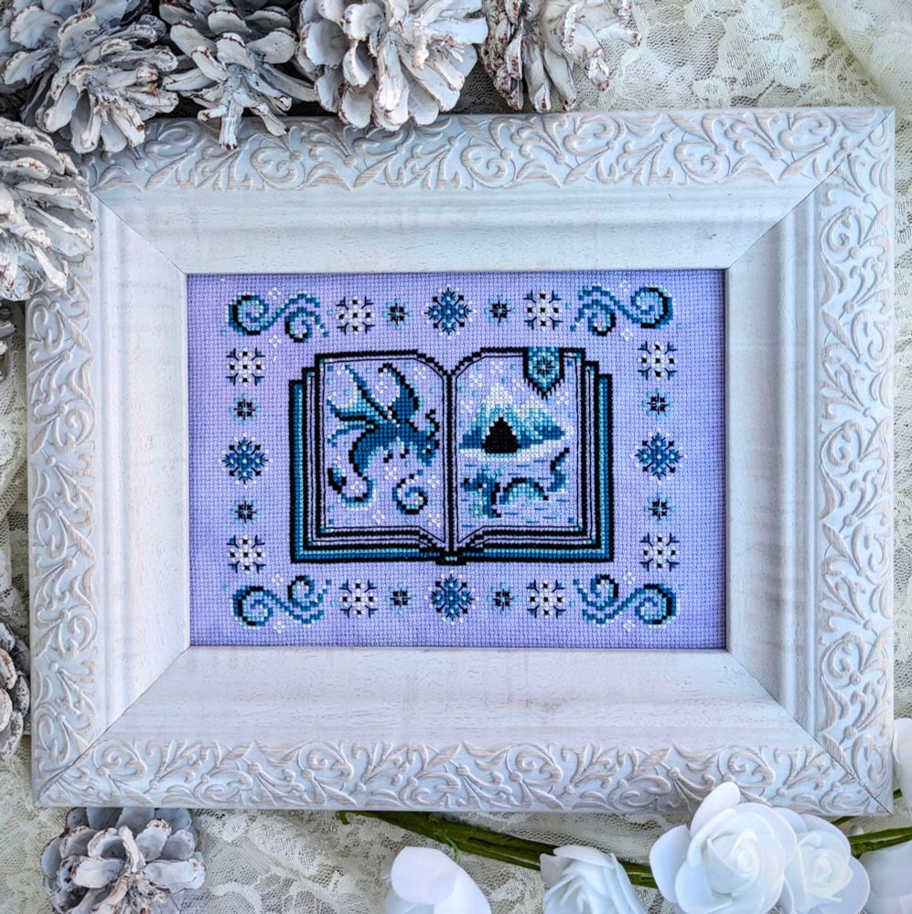 A finished cross stitch pattern features a blue open faced book with a blue dragon on the left page and a mountain and sea monster on the right page. Snowflakes surround the pattern border.