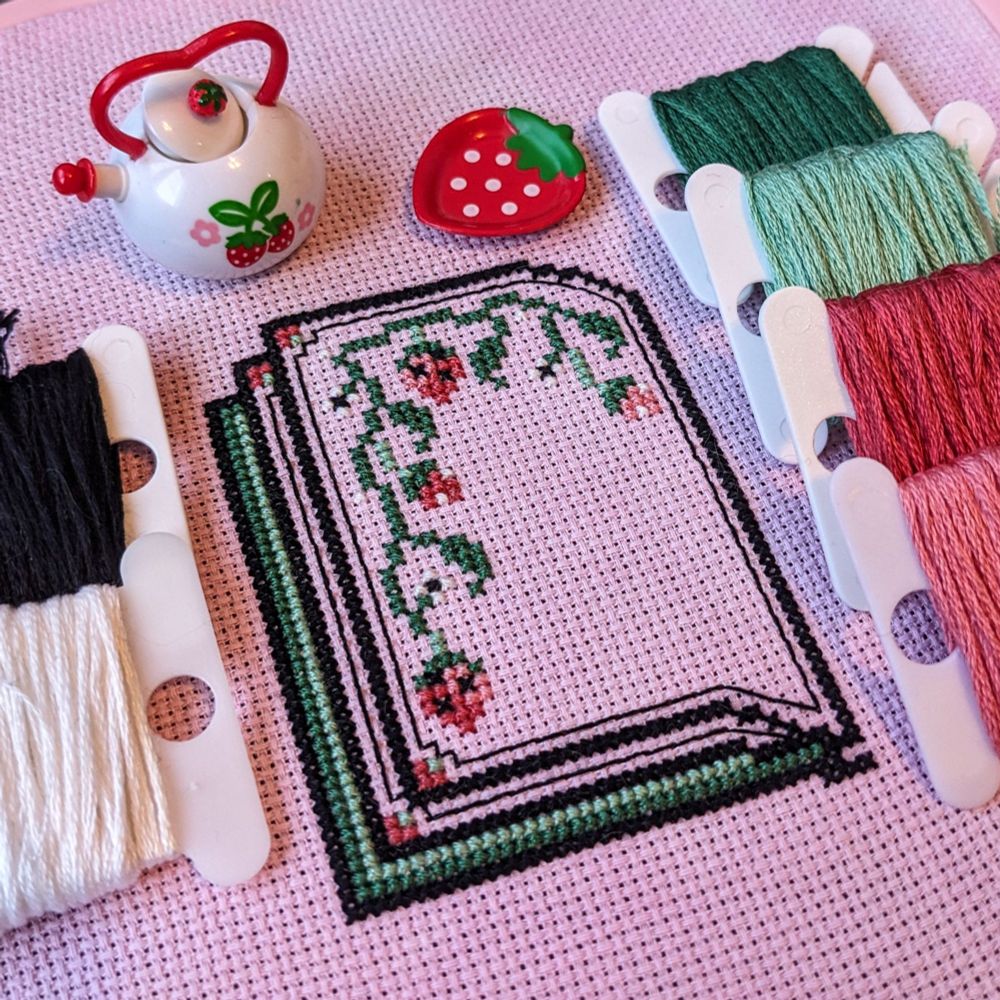 A sneak peek of a cross stitch pattern designs. Shows half an open book with strawberries on the book page. A little strawberry tea pot and plate are shown for decoration