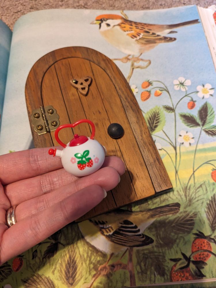 A hand holding a miniature strawberry themed teapot and showing a fairy door behind