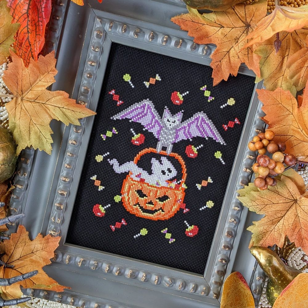A cross stitch pattern features a spooky cute bat and ghost kitty floating in the air to steal Halloween candy 