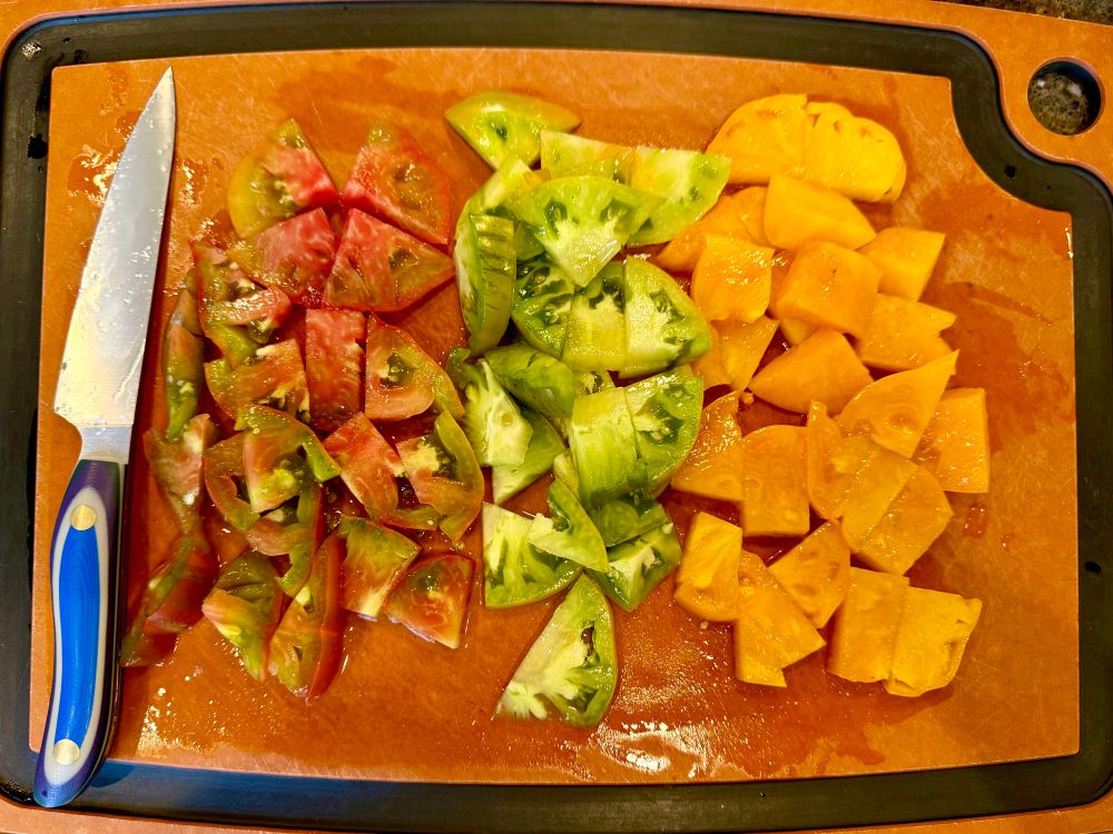 A cutting board with large pieces of cut tomato in three varieties starting with a row of red and green tie-dye, a row of all green (intentionally, not unripe), & a row of meaty golden fruits. 