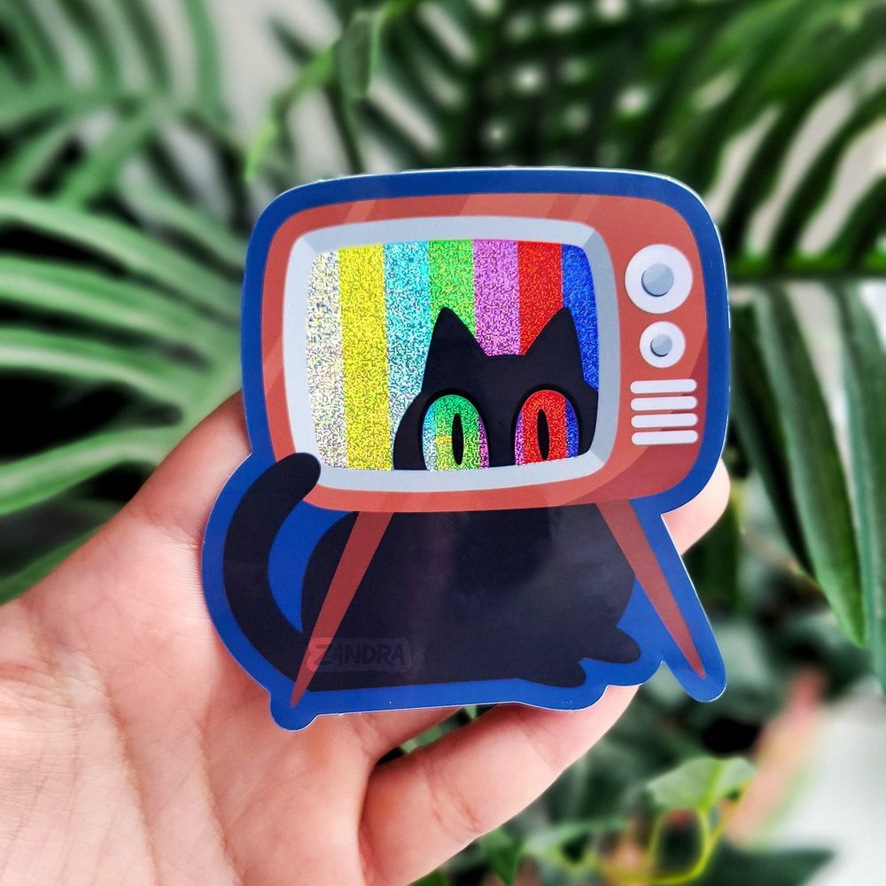 a photo of a sticker design by Zandra Illustration featuring an old-looking brown television with a black cat. The black cat is peering through the television screen from behind and colorful broadcast bars are shown on the screen. The sticker has a static-y glitter effect on both the tv screen and the black cat's eyes, as if the television signal is impacting the cat's vision.