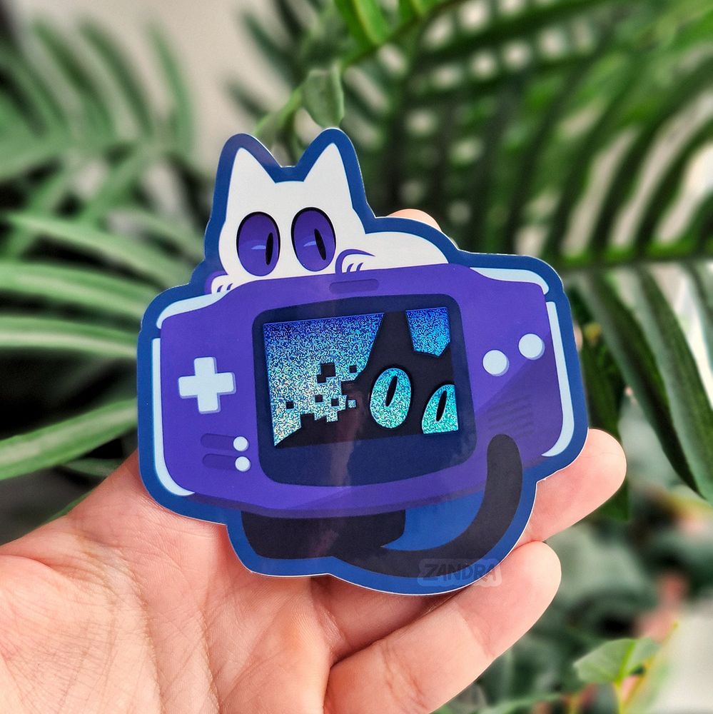 a photo of a sticker design by Zandra Illustration featuring a popular handheld game console. A black cat is peering through the blue screen of the console from behind, and it's eyes are the same color of glittery blue as the screen. A white cat sits on top of the console peering down.