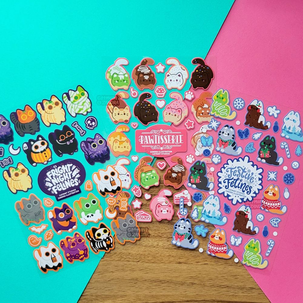a photo of three cat-themed sticker sheets with clear backgrounds on a backdrop of colorful photo paper. Each sheet has a theme of halloween, baked goods, and winter respectively.